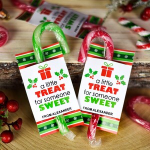 Pu&ograve; includere: Due bastoncini di zucchero con involucri a strisce rosse e verdi festivi sono legati con un nastro rosso e verde a un'etichetta regalo bianca. L'etichetta recita "un piccolo regalo per qualcuno dolce da parte di Alexander".