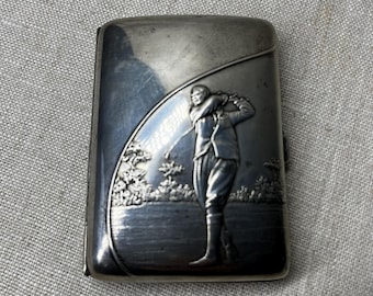 Könnte beinhalten: Silbernes Zigarettenetui mit einem geprägten Design eines Golfers, der einen Schläger schwingt. Das Etui hat einen Klappdeckel und eine glatte, polierte Oberfläche.