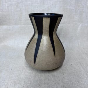May include: A ceramic vase with a black rim and vertical black stripes against a beige background. The vase has a rounded body and a slightly flared neck, creating a visually appealing shape. The design is simple yet elegant.