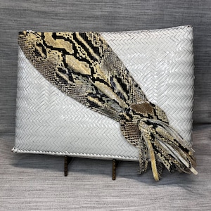 May include: A white woven clutch with a decorative flap made of snakeskin-patterned material. The flap is tan, brown, and black. The clutch is rectangular and sits on a small, brown metal stand.