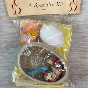 May include: A Specialty Kit in a clear plastic bag. The kit includes a pink figurine with a yellow hat, a white foam egg, and an egg-shaped basket filled with flowers and a blue ribbon. The packaging has a cream-colored label.