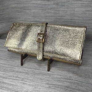 May include: A gold-colored clutch with a textured finish. The clutch features a buckle closure and a strap. The accessory is displayed on a small, brown metal stand.