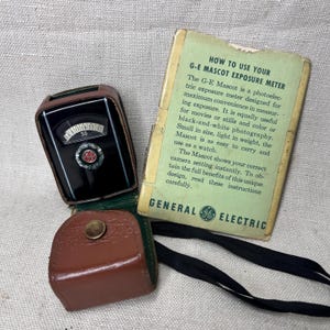 May include: Vintage G-E Mascot exposure meter with a black and silver face, housed in a brown leather case. Includes a green and yellow instruction booklet with the text "HOW TO USE YOUR G-E MASCOT EXPOSURE METER". A black strap is also visible.