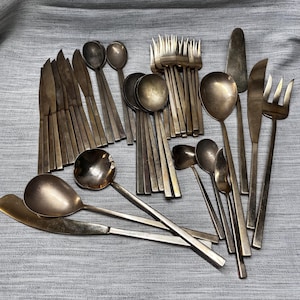 May include: A collection of vintage silverware, including knives, forks, spoons, and serving utensils. The silverware has a tarnished, antique finish, with a variety of spoon sizes and fork tines. The arrangement is on a gray fabric surface.