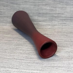 May include: A maroon-colored, hourglass-shaped vase. The vase has a wide base and a narrow middle, with a flared opening at the top. The color transitions from a darker shade at the top to a lighter shade at the base.