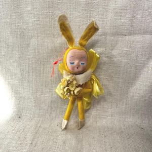 May include: A yellow felt bunny doll with closed eyes and a white fur collar holds a bouquet of yellow felt flowers.