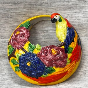 May include: A ceramic wall hanging featuring a colorful parrot perched on a branch with flowers. The parrot has yellow, red, and blue feathers. The flowers are in shades of red and blue, set against a yellow background. The words "MADE IN JAPAN" are visible.
