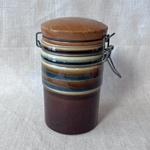 May include: A ceramic canister with a wooden lid and metal clasp. The canister features horizontal bands of brown, blue, and beige. The lid is a light brown color. The metal clasp is silver.