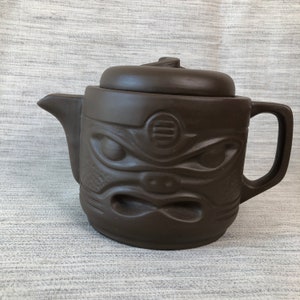 May include: A brown ceramic teapot with a lid and handle. The teapot features a carved face design on the front, with detailed eyes, nose, and mouth. The teapot is designed for brewing tea.