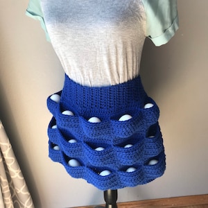 May include: A blue crocheted skirt with white balls in the pockets. The skirt is on a black mannequin.
