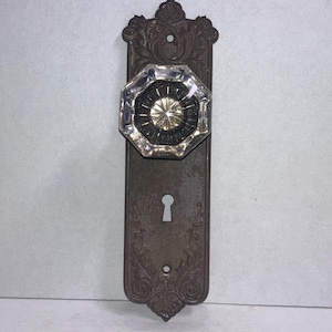 May include: An octagonal clear glass door knob attached to a rectangular dark brown metal backplate. The backplate features an ornate embossed floral pattern and a central keyhole. The glass knob has a faceted design with a starburst center.