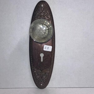 May include: An oval-shaped, dark bronze-toned metal door plate featuring intricate embossed floral patterns, a central clear glass knob with a faceted design, and a keyhole opening. A small white rectangular sticker with the letters BB is attached.