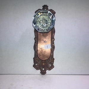 May include: An octagonal clear glass doorknob attached to a rectangular copper-toned metal backplate with ornate decorative edges. The metal plate features a small white sticker with the handwritten number 1 centered on its surface.