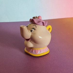 May include: A pink and yellow ceramic teapot figurine with a purple lid and a smiling face. The teapot has a spout and a handle.