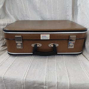 Pode incluir: Uma mala vintage castanha com uma pega preta e fechos prateados. A mala tem uma moldura branca e preta e uma pequena etiqueta que diz "British Airways".
