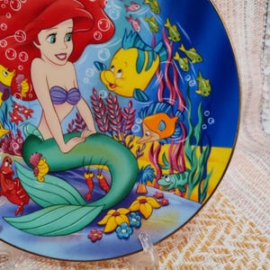 Puede incluir: Un plato decorativo con una colorida ilustraci&oacute;n de Ariel, la Sirenita, y sus amigos acu&aacute;ticos. El plato presenta una escena submarina azul y amarilla con peces e hipocampos. El plato est&aacute; sostenido por un soporte de pl&aacute;stico transparente.