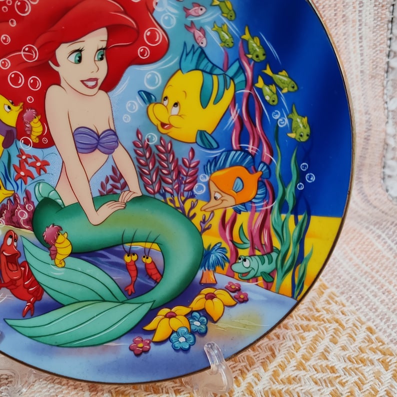 Puede incluir: Un plato decorativo con una colorida ilustraci&oacute;n de Ariel, la Sirenita, y sus amigos acu&aacute;ticos. El plato presenta una escena submarina azul y amarilla con peces e hipocampos. El plato est&aacute; sostenido por un soporte de pl&aacute;stico transparente.