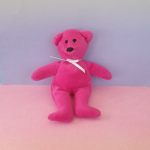 May include: A plush, cerise teddy bear with a white ribbon tied around its neck. The bear has black button eyes and a black stitched mouth. The background is a gradient of pink and purple.