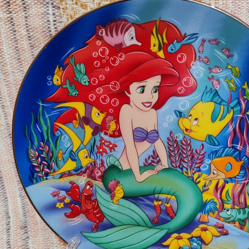 Puede incluir: Un plato decorativo con Ariel de La Sirenita de Disney. El plato tiene un fondo azul con Ariel, cabello rojo, un top de concha p&uacute;rpura y una cola verde. Peces coloridos y otras criaturas marinas la rodean.