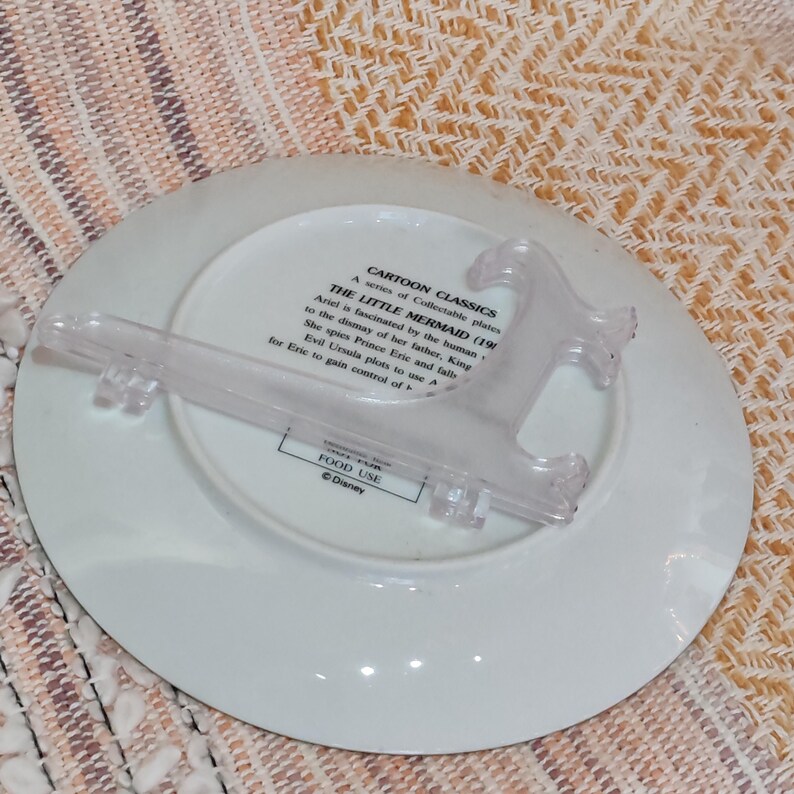 Puede incluir: Un plato de cer&aacute;mica blanca de la serie Cartoon Classics, con texto sobre La Sirenita. Un soporte de pl&aacute;stico transparente para platos est&aacute; adjunto. El plato est&aacute; marcado "FOOD USE" y "&copy;Disney."