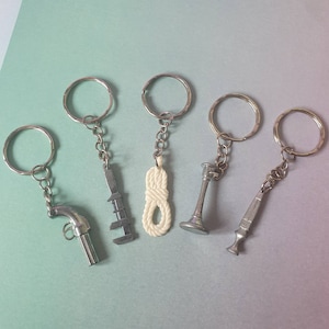 May include: A collection of five keychains with silver-colored metal rings and chains. The charms include a pistol, a wrench, a rope knot, a candlestick, and a decorative object. The background is a gradient of blue and green.