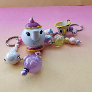 Pode incluir: Uma coleção de chaveiros com personagens de desenhos animados. Os chaveiros incluem uma chaleira branca com tampa roxa, uma xícara amarela e várias contas iridescentes. Os chaveiros são conectados com anéis de metal prateado.