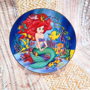 Puede incluir: Un plato decorativo con Ariel de La Sirenita. El plato tiene un fondo azul con ilustraciones coloridas de Ariel, peces y plantas submarinas. Ariel tiene el pelo rojo y una cola verde. El plato se exhibe en un soporte transparente.