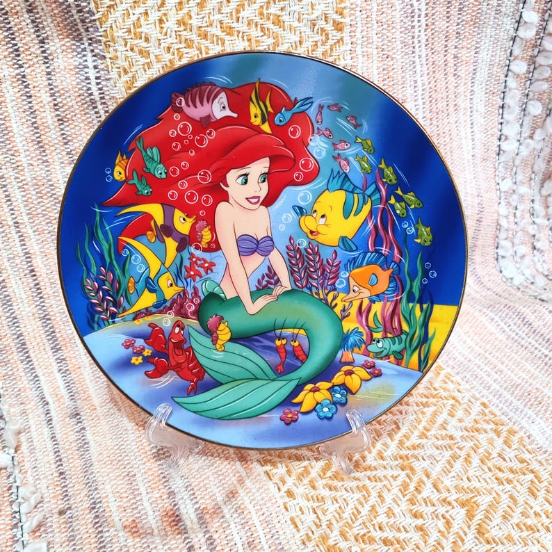 Puede incluir: Un plato decorativo con Ariel de La Sirenita. El plato tiene un fondo azul con ilustraciones coloridas de Ariel, peces y plantas submarinas. Ariel tiene el pelo rojo y una cola verde. El plato se exhibe en un soporte transparente.