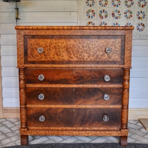 May include: A wooden chest of drawers with a warm, reddish-brown finish. The chest features four drawers with crystal knobs and a top section with a decorative burl wood pattern. The piece has a classic, antique design.