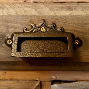 May include: A decorative drawer pull with a rectangular opening and ornate details. The pull is made of metal and has a dark brown finish.