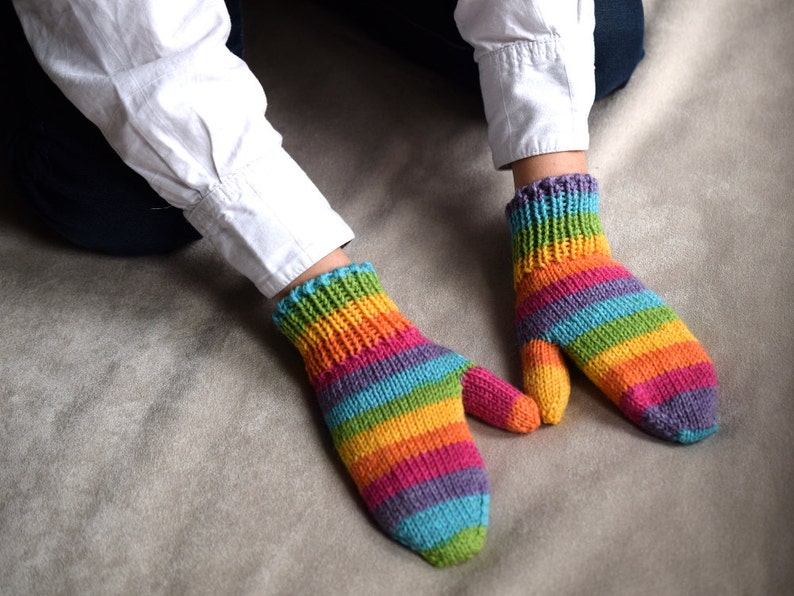 Rainbow colored wool mittens presented on child's hands. Rainbow stripes are mismatched on the two mittens. Color progression is: green, blue, purple, magenta, orange, yellow repeating.