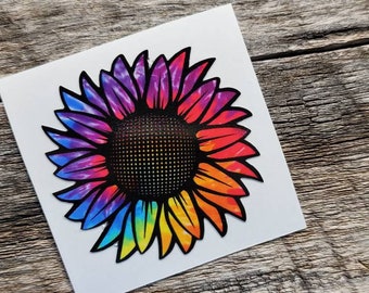 May include: A colorful sunflower sticker with a tie-dye effect. The petals transition from purple to red, orange, yellow, and green. The center is black with a dotted pattern. The sticker is on a white square.