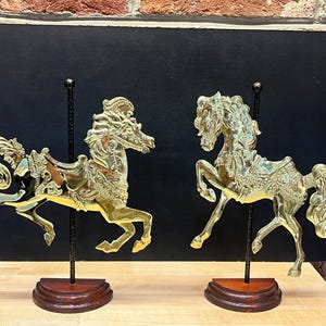 May include: Two polished brass carousel horse figurines, each mounted on a thin black vertical rod and a semi-circular dark wood base. The horses feature intricate embossed saddles and flowing manes, captured in a dynamic galloping pose.