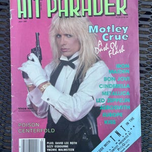 May include: A July 1987 issue of Hit Parader magazine featuring a cover photograph of Vince Neil with long blonde hair, wearing a white shirt, black vest, and bow tie, holding a black revolver while wearing white gloves against a pink background.