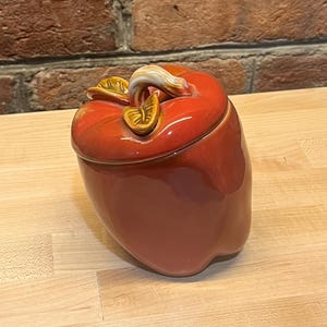May include: A glossy red ceramic jar shaped like a bell pepper with a removable lid. The lid features a sculpted stem and two small leaves in shades of brown and cream. The body of the jar has a smooth, rounded form with a vibrant, reflective finish.