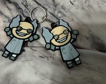 Puede incluir: Dos llaveros de goma idénticos que representan a un científico de dibujos animados con pelo gris, gafas redondas y bata blanca. Las figuras tienen una amplia sonrisa y están unidas a una anilla de metal plateado con una cadena.