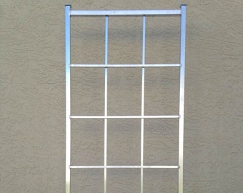 May include: A silver metal trellis with a rectangular shape and a grid pattern. The trellis is standing upright on a grassy surface.