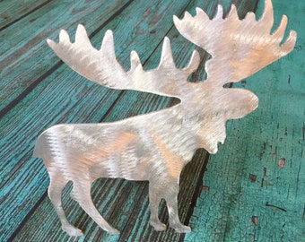 May include: A silver metal moose silhouette with a brushed finish. The moose is standing on a teal blue wooden surface.