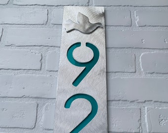May include: A silver metal house number sign with a teal blue background. The numbers 927 are displayed in a vertical line with a dove silhouette at the top.