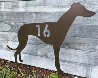 May include: A brown metal silhouette of a greyhound with the number 16 cut out of the side. The silhouette is mounted on two metal stakes and is standing in front of a gray wooden fence.