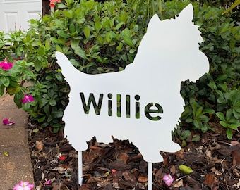 May include: White metal silhouette of a dog with the name "Willie" cut out. The silhouette is standing on a white stake in a garden setting.