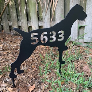 May include: Black metal silhouette of a dog with the house number 5633 cut out. The silhouette is mounted on a metal stake and is placed in the ground.