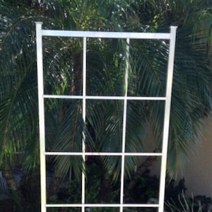 May include: A silver metal trellis with a square grid pattern. The trellis is standing in a garden with brown mulch and green plants.