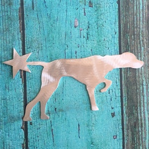 May include: A silver metal silhouette of a dog with a star on its tail. The dog is standing on a turquoise wooden background.