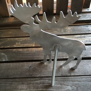 May include: A silver metal moose silhouette with large antlers. The moose is standing on a metal rod that is inserted into the ground.