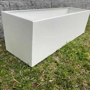 Puede incluir: Un macetero rectangular blanco hecho de un material liso. El macetero está sentado en un lecho de hierba verde.