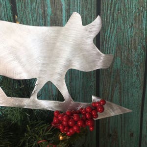 May include: A silver metal pig silhouette Christmas tree topper with a red berry sprig attached to the arrow.