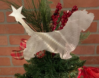 May include: A silver metal Christmas tree topper shaped like a dog with a star on its head. The topper is on a green Christmas tree with red and white ribbon.