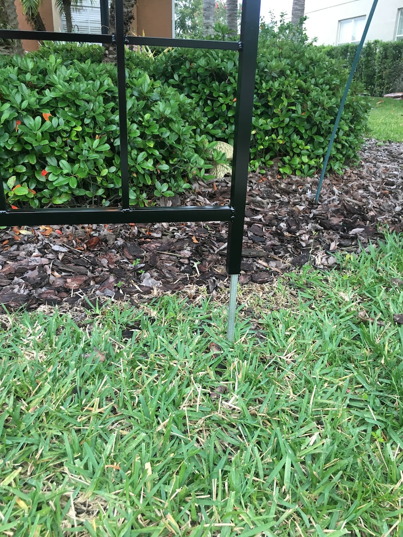 May include: A black metal garden trellis with a square design. The trellis is anchored to the ground with a metal stake.