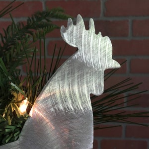 Puede incluir: Un adorno de gallo de metal plateado en forma de silueta para un &aacute;rbol de Navidad. El gallo est&aacute; hecho de metal y tiene un acabado cepillado.